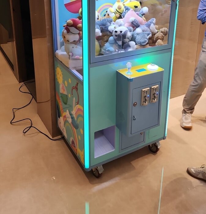 室內場地上一部淺藍色機身、彩虹圖案的夾公仔機,下櫃有兩個投幣口與出獎口,底座有轆,一條黑色電線由機身後方沿地面伸向牆邊