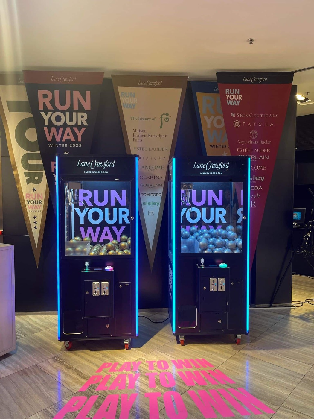 商場活動中一對黑色夾公仔機，機身與玻璃印有白色 RUN YOUR WAY 字樣，機身邊緣亮起藍色燈條，地上投射粉紅色字句，背景懸掛三角旗