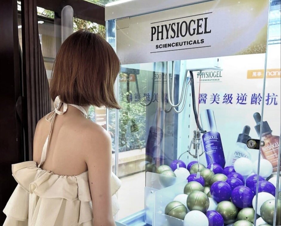 一部透明機身的夾公仔機，頂牌印有護膚品牌標誌，機內背板印有產品圖片及中文宣傳字，機內放滿紫色、金色及白色膠球，一位女士正在按操作台的按鈕