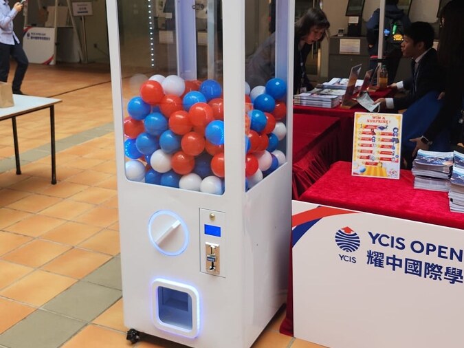 白色方扭蛋機放在學校開放日的攤位旁,機內裝滿紅、藍、白三色扭蛋,旁邊桌上放著換領告示牌