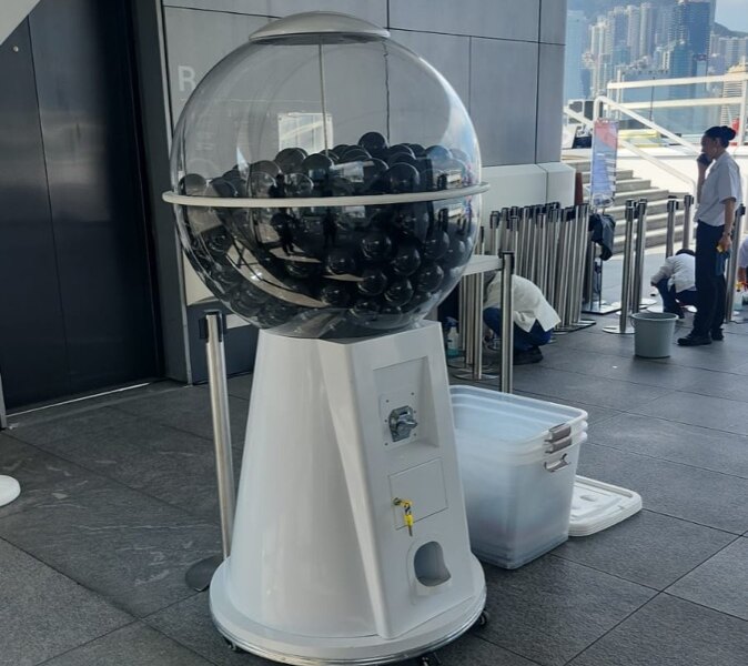 戶外平台的升降機口旁放著一部白色底座的大圓扭蛋機,透明圓球內裝滿黑色扭蛋,底座有轆,四周有伸縮圍欄,遠處可見山景與高樓