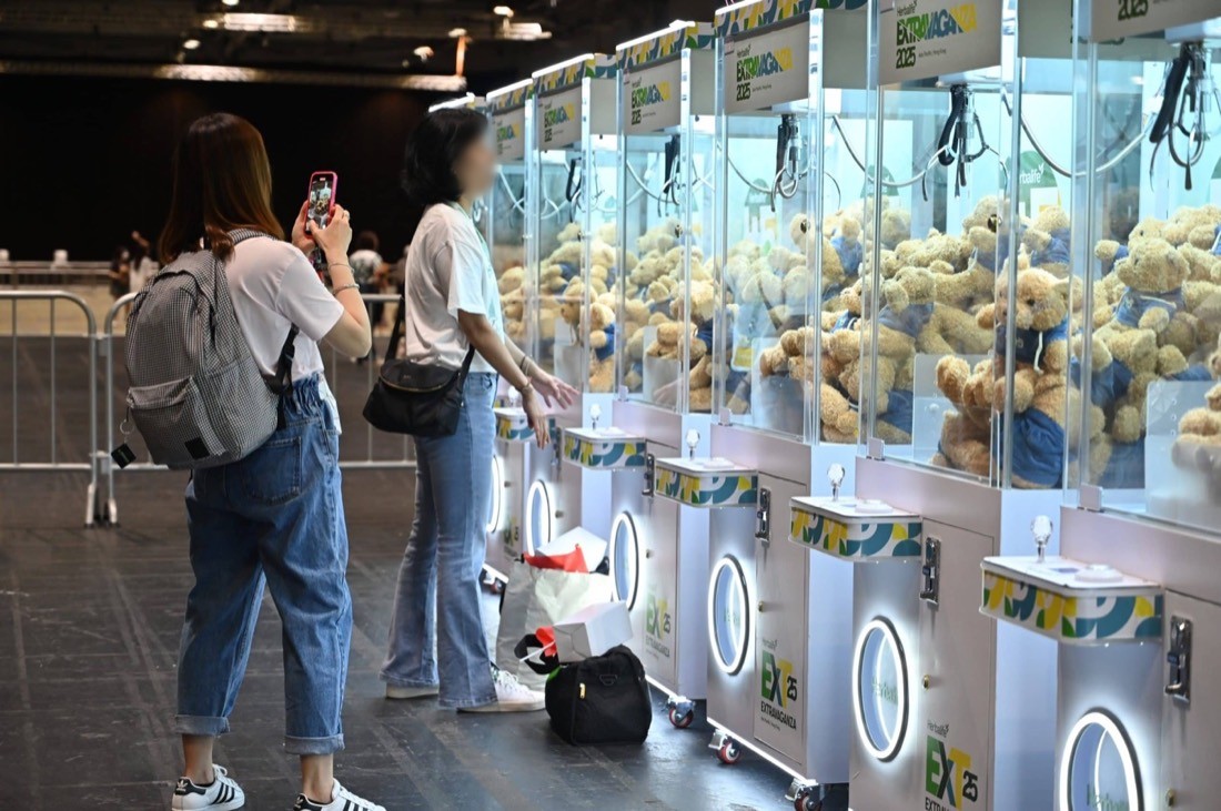 商場活動現場一排品牌夾公仔機，每部機內堆滿同一款穿藍色上衣的米色毛公仔，兩名參觀者在機前拍照